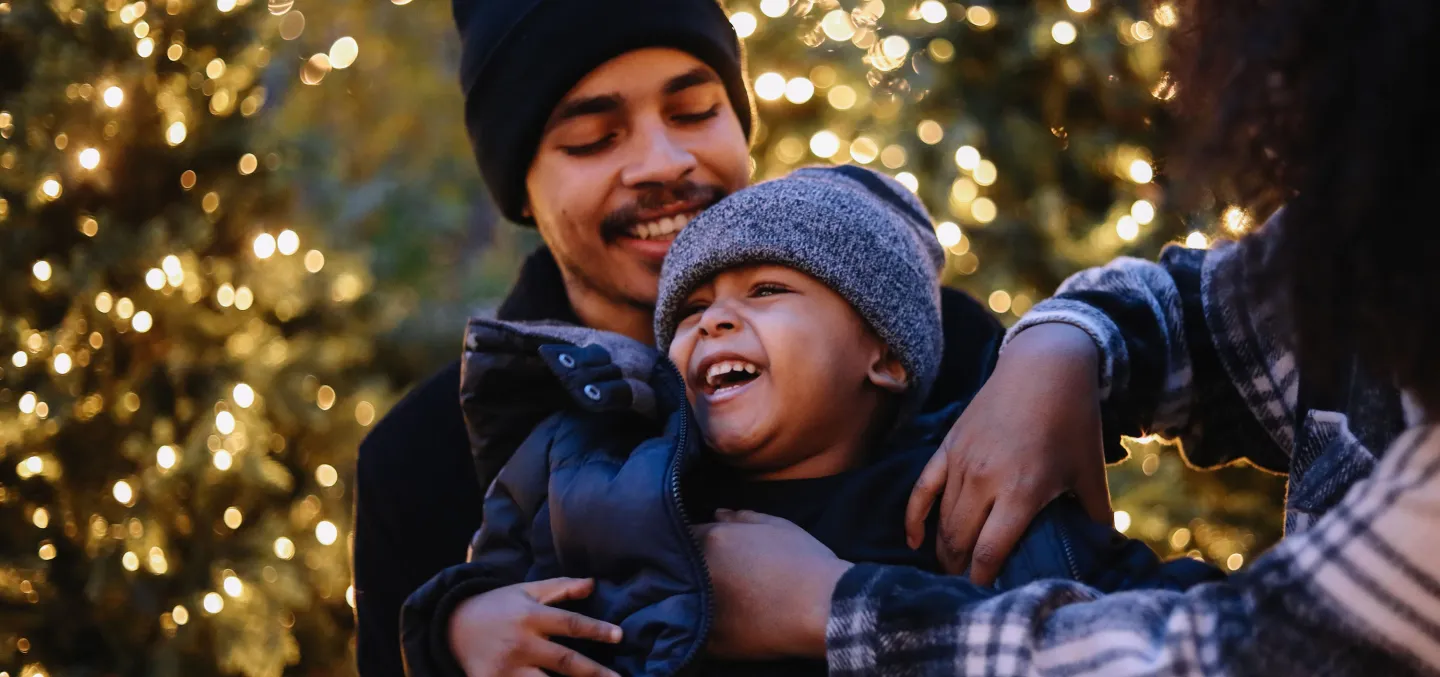 Father holding laughing child in winter clothes with holiday lights glowing in the background.