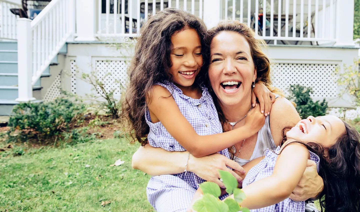 Mother laughing with her two young daughters in matching dresses while playing together outside a white house.