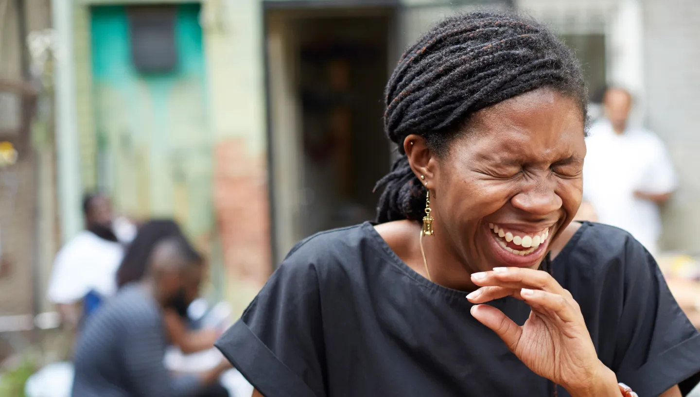 Woman laughing joyfully at an outdoor gathering, surrounded by people in a relaxed, urban backyard setting.