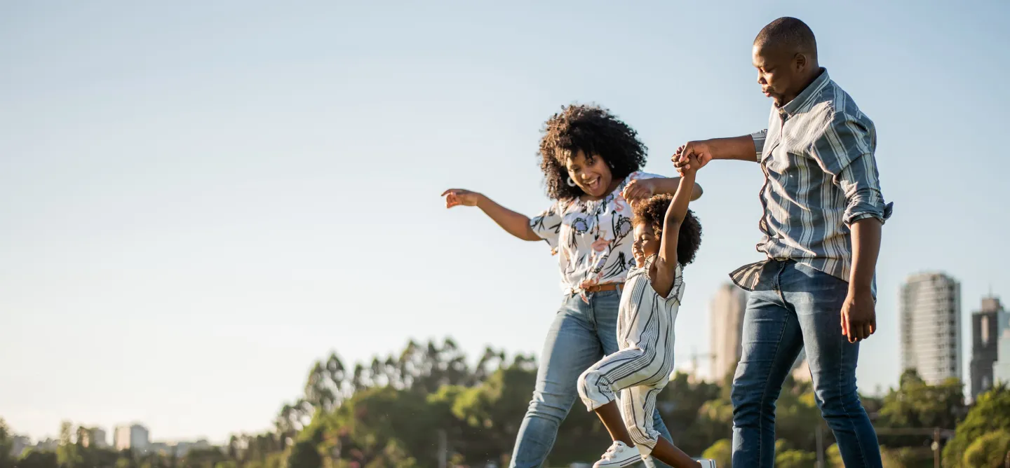 Parents swing their young daughter by the arms while walking together in a park on a sunny day.