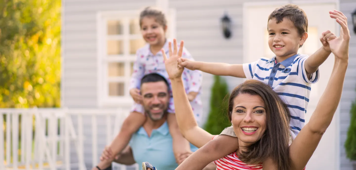 Smiling parents give their young children piggyback rides outside their home on a sunny day.