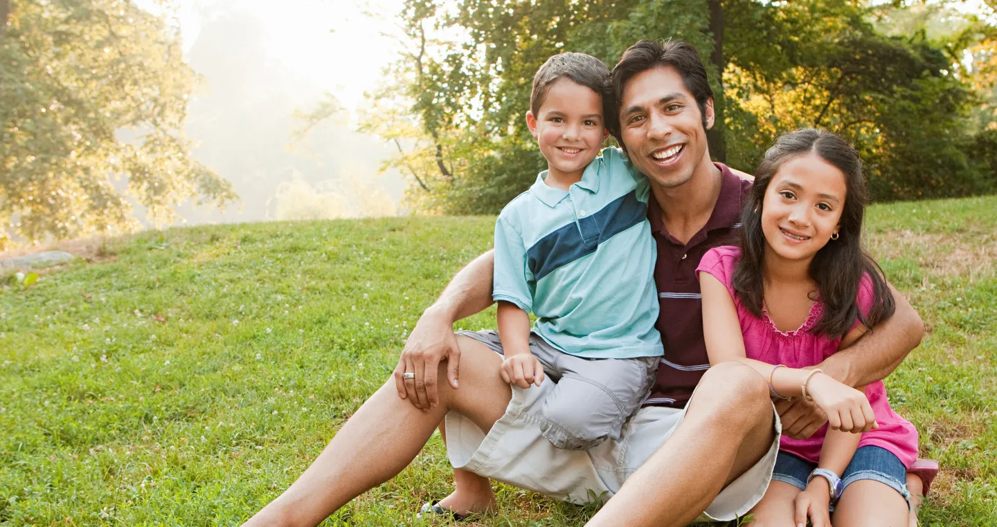 Smiling father sits on the grass with his young son and daughter on a sunny day in the park.