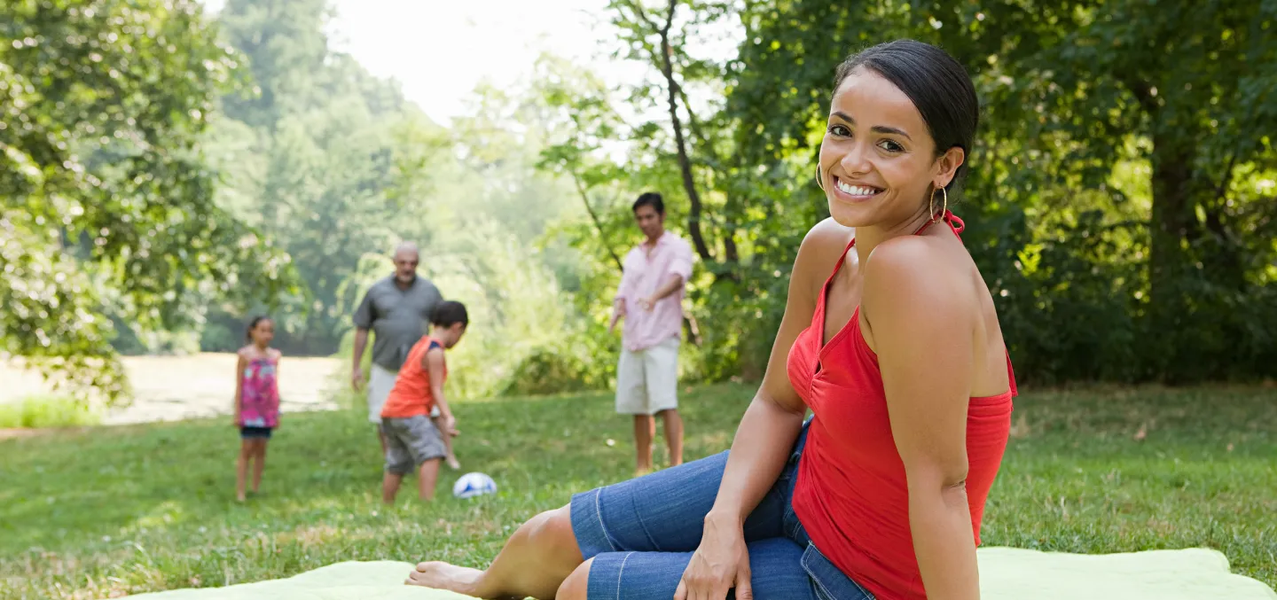 Woman in red top smiles while sitting on a picnic blanket as family plays in the background at a sunny park.