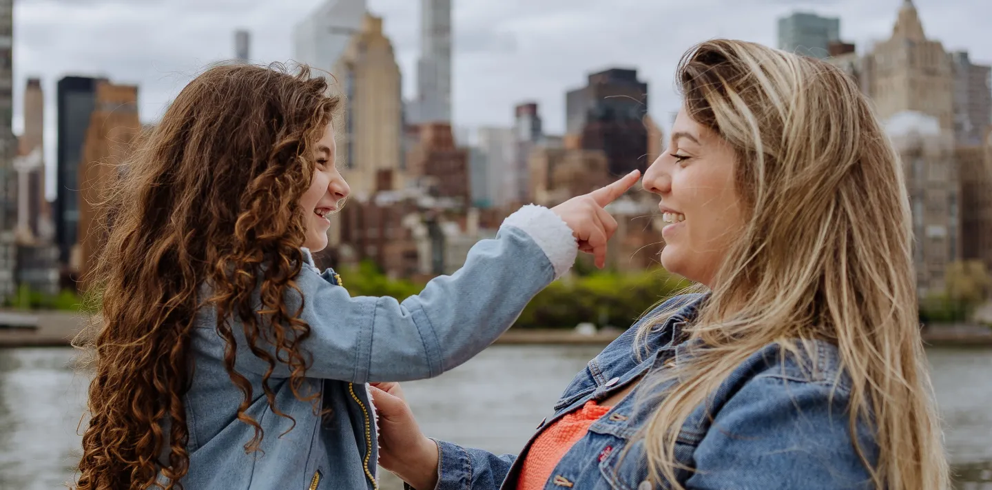Smiling young girl touches her mother’s nose playfully as they enjoy time together with a city skyline in the background.
