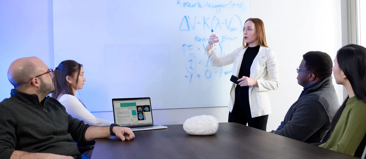 A woman in a white blazer presents brain data on a whiteboard to a small group of seated colleagues.