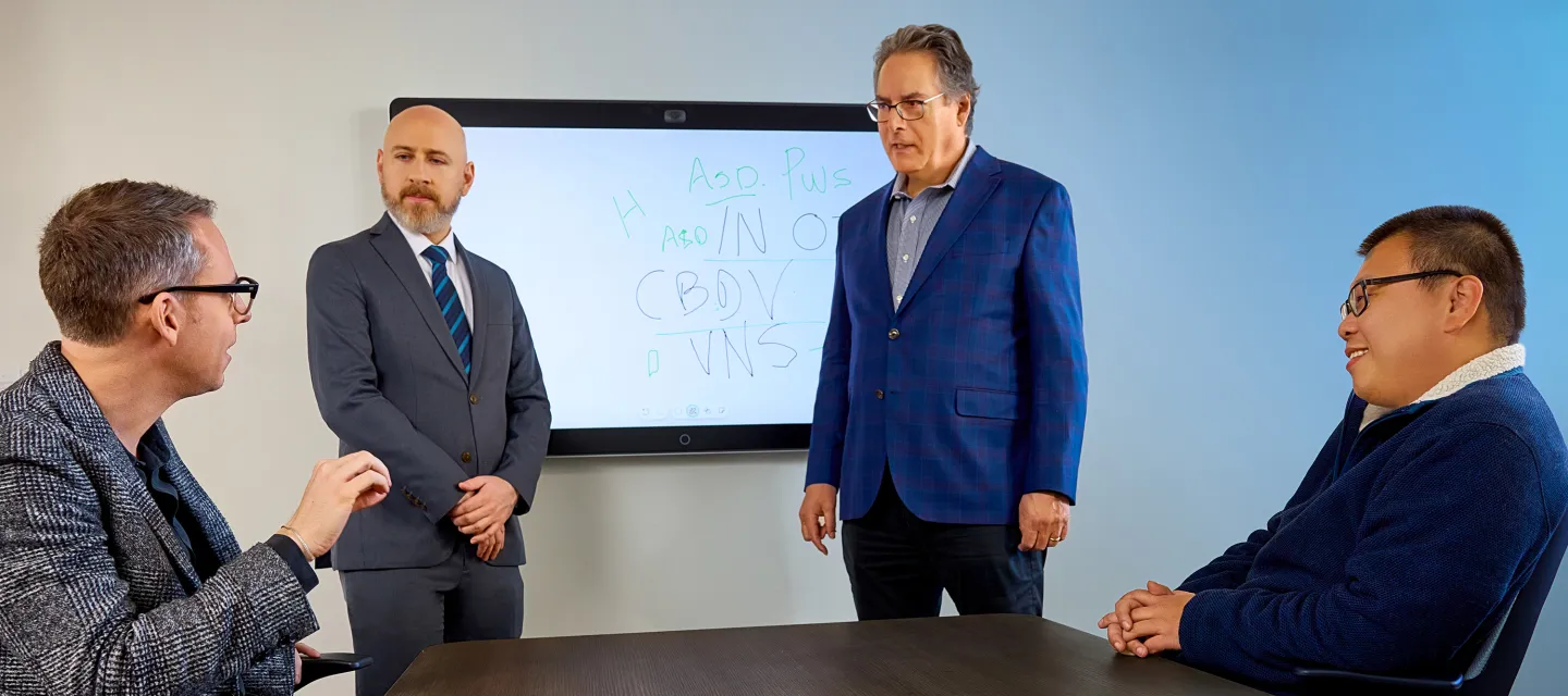 Four men in a conference room discuss in front of a whiteboard with handwritten notes and acronyms.