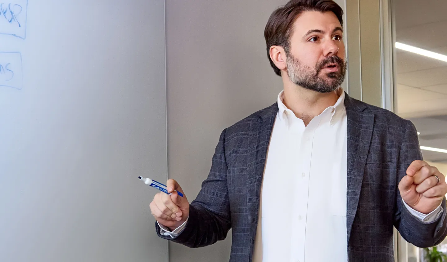 Man in blazer holding marker, explaining at whiteboard in modern office setting.