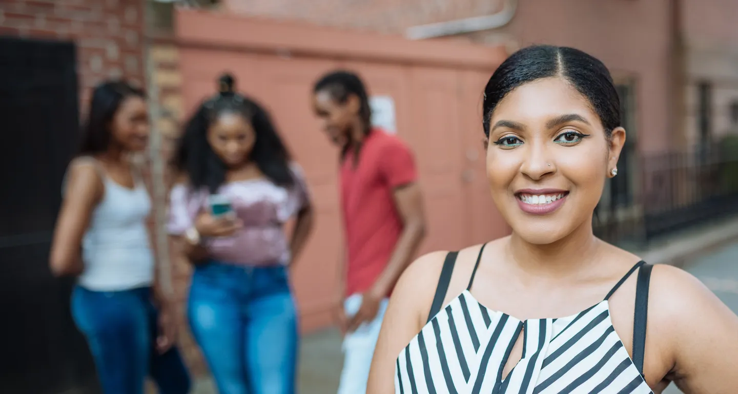 Smiling young woman in striped top stands in focus while three friends chat in the blurred background on a city street.