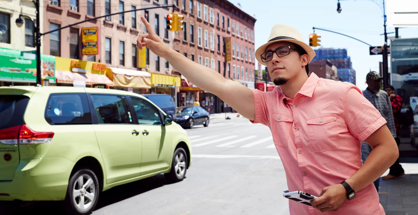 Man in a pink shirt and hat hails a cab on a busy city street while holding his phone.