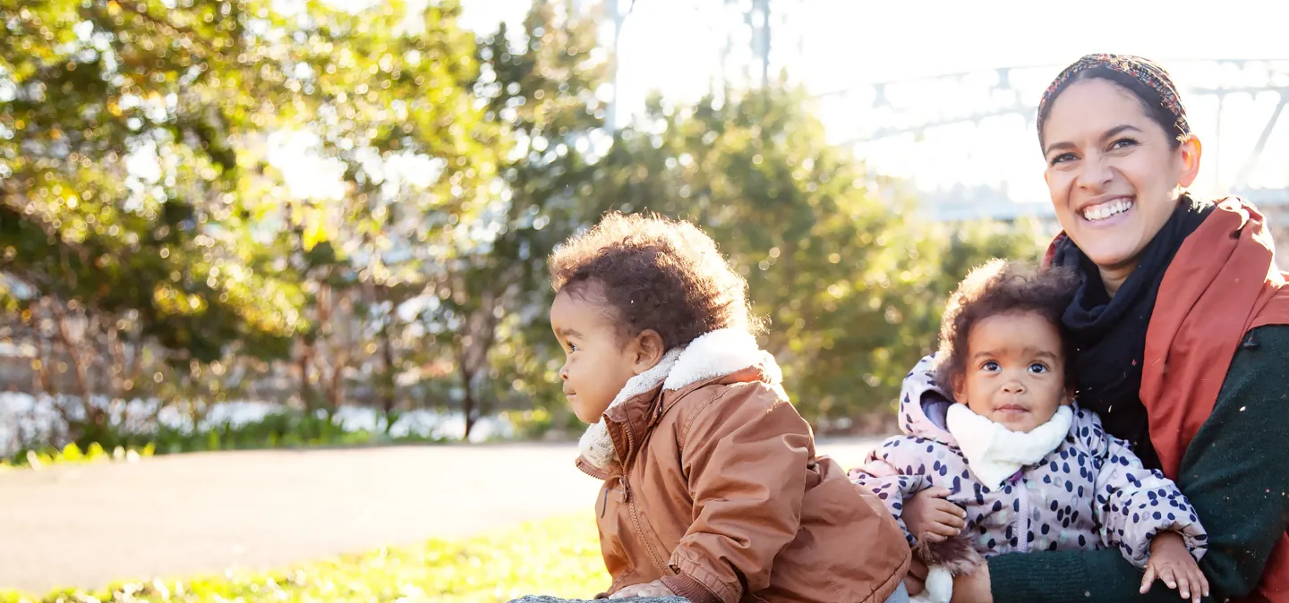 Smiling mother sits outdoors holding a baby while another young child plays nearby in warm jackets on a sunny day.