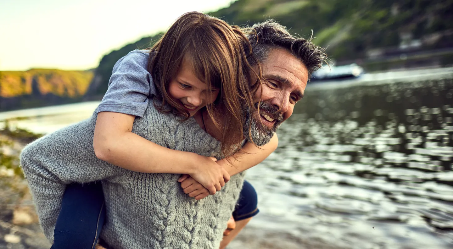 Smiling child rides on man's back by a scenic riverbank