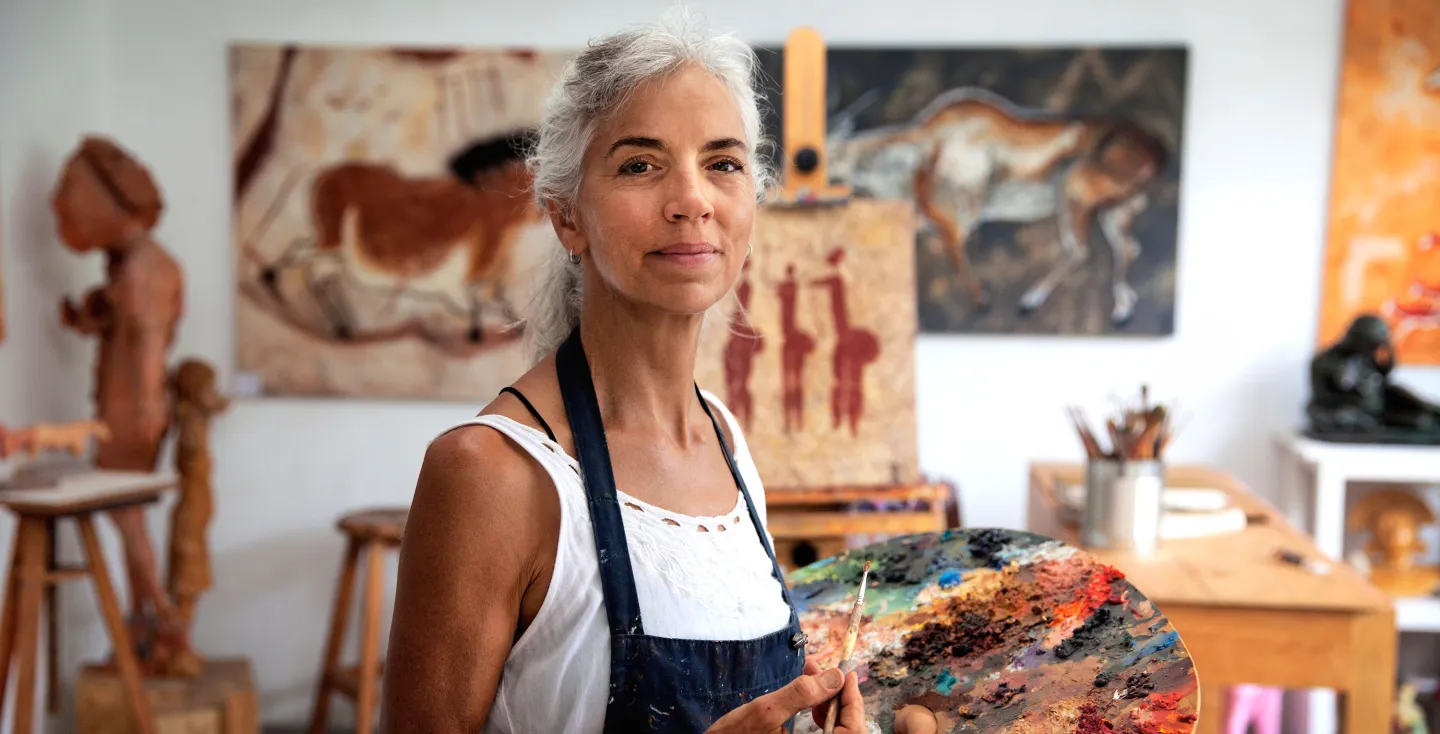 Older woman artist holding paintbrush and palette in a studio with abstract paintings