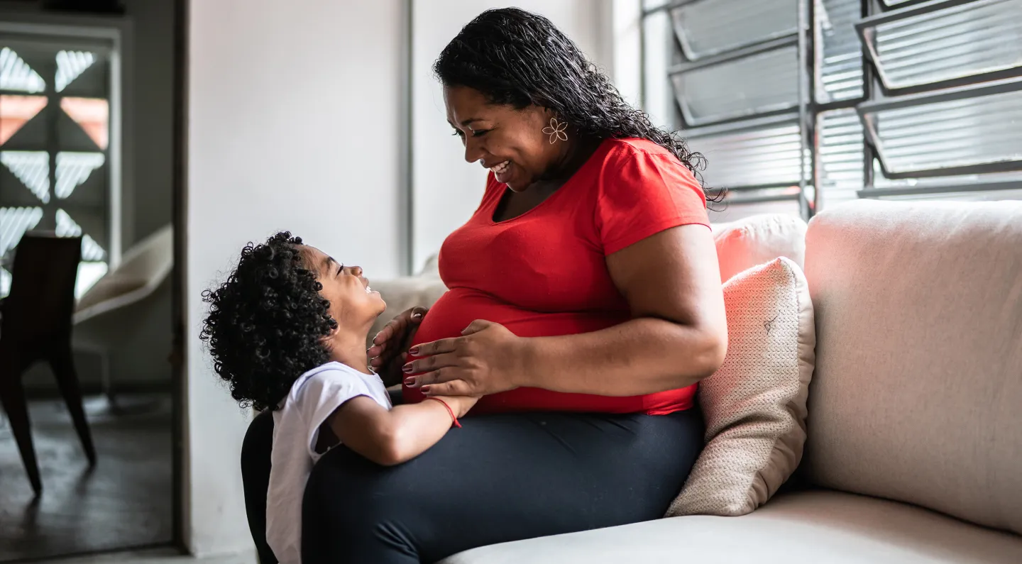 Pregnant woman smiles at young child while sitting on a couch in a sunlit living room.