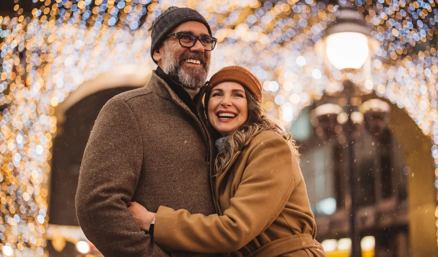 Smiling couple in winter coats hugging under festive holiday lights on a snowy evening