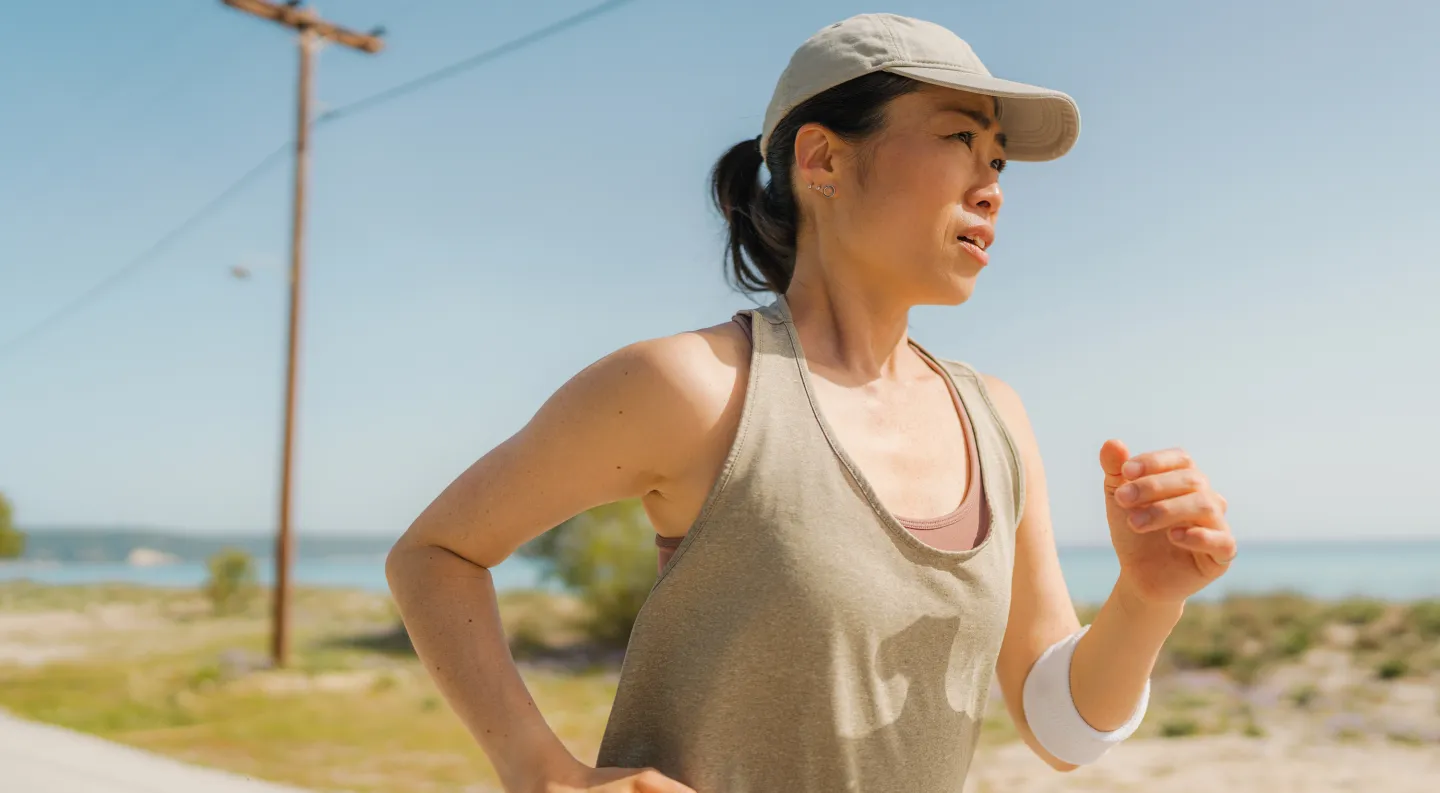 Asian woman running outdoors in sunny weather wearing a cap and athletic gear.