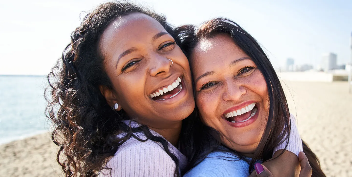 Two women smiling and hugging each other on a sunny beach, with buildings and palm trees in the background