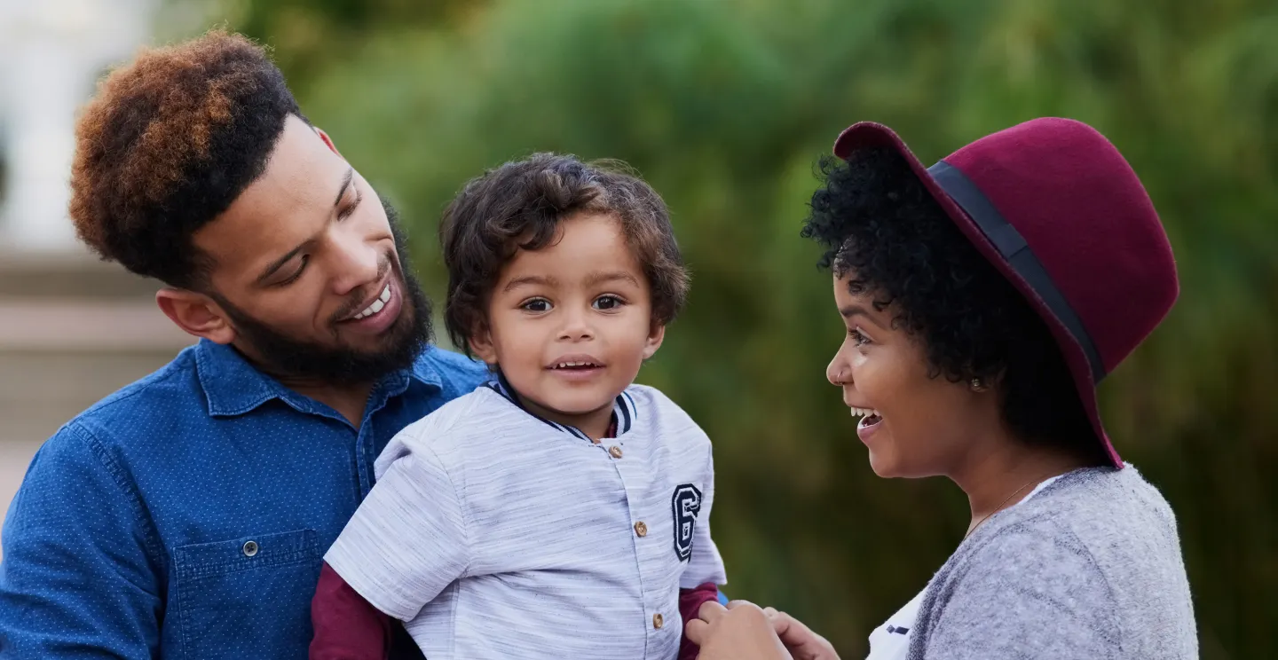 Smiling parents enjoy a moment with their young child outdoors on a sunny day.