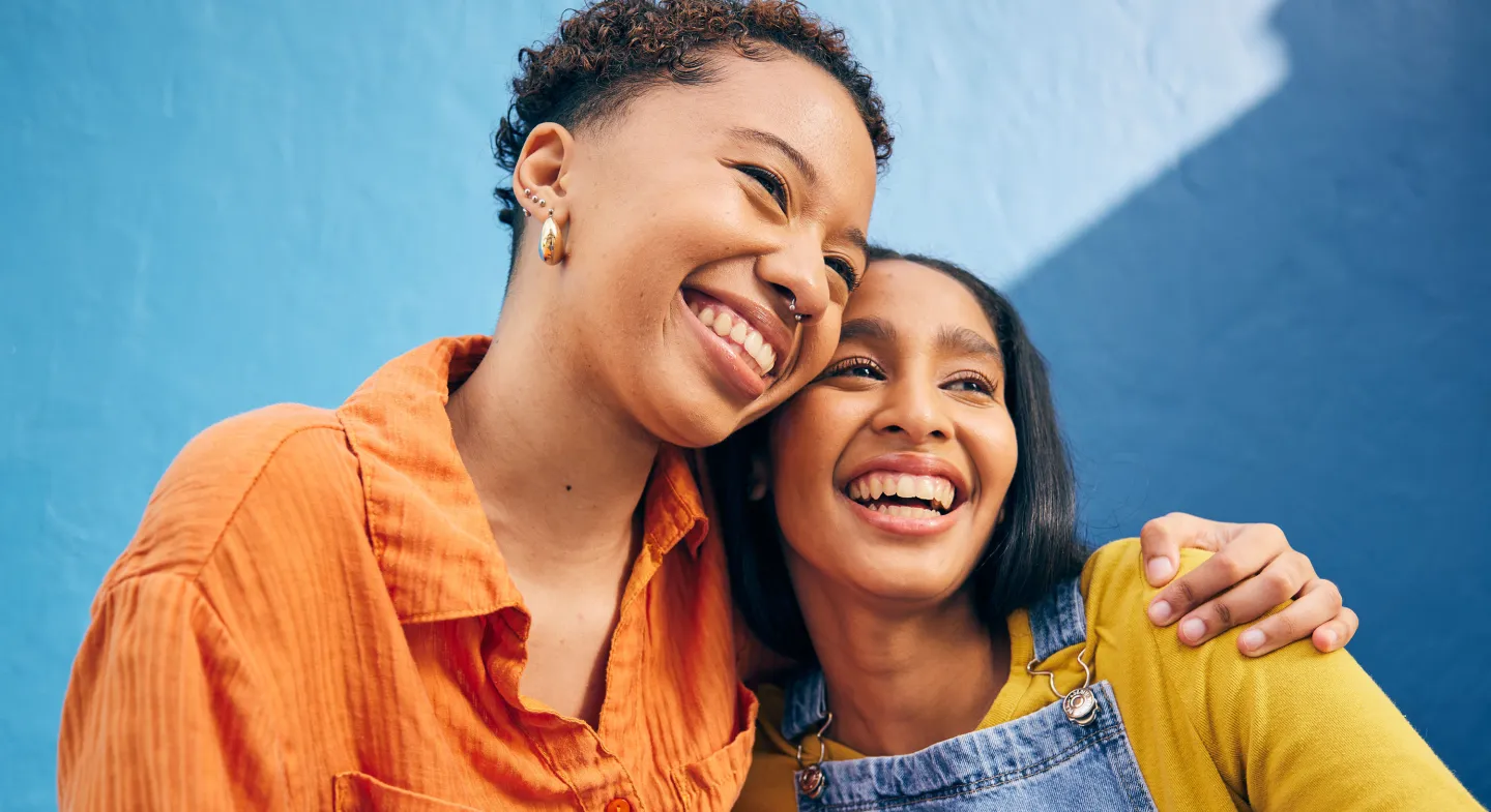 Two friends smiling and hugging in front of a bright blue wall on a sunny day