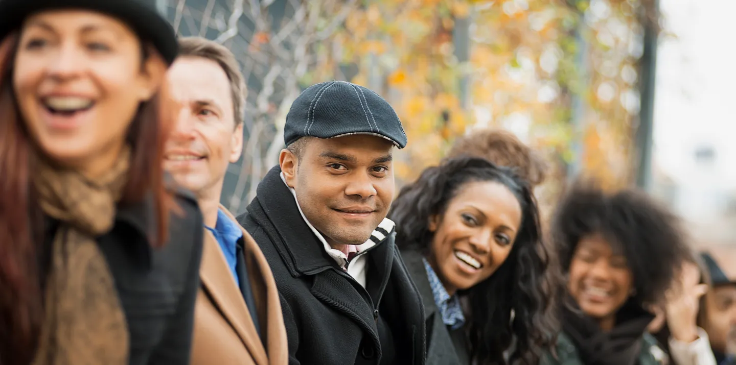 Man in black cap smiles among a diverse group of people standing outdoors in fall weather.