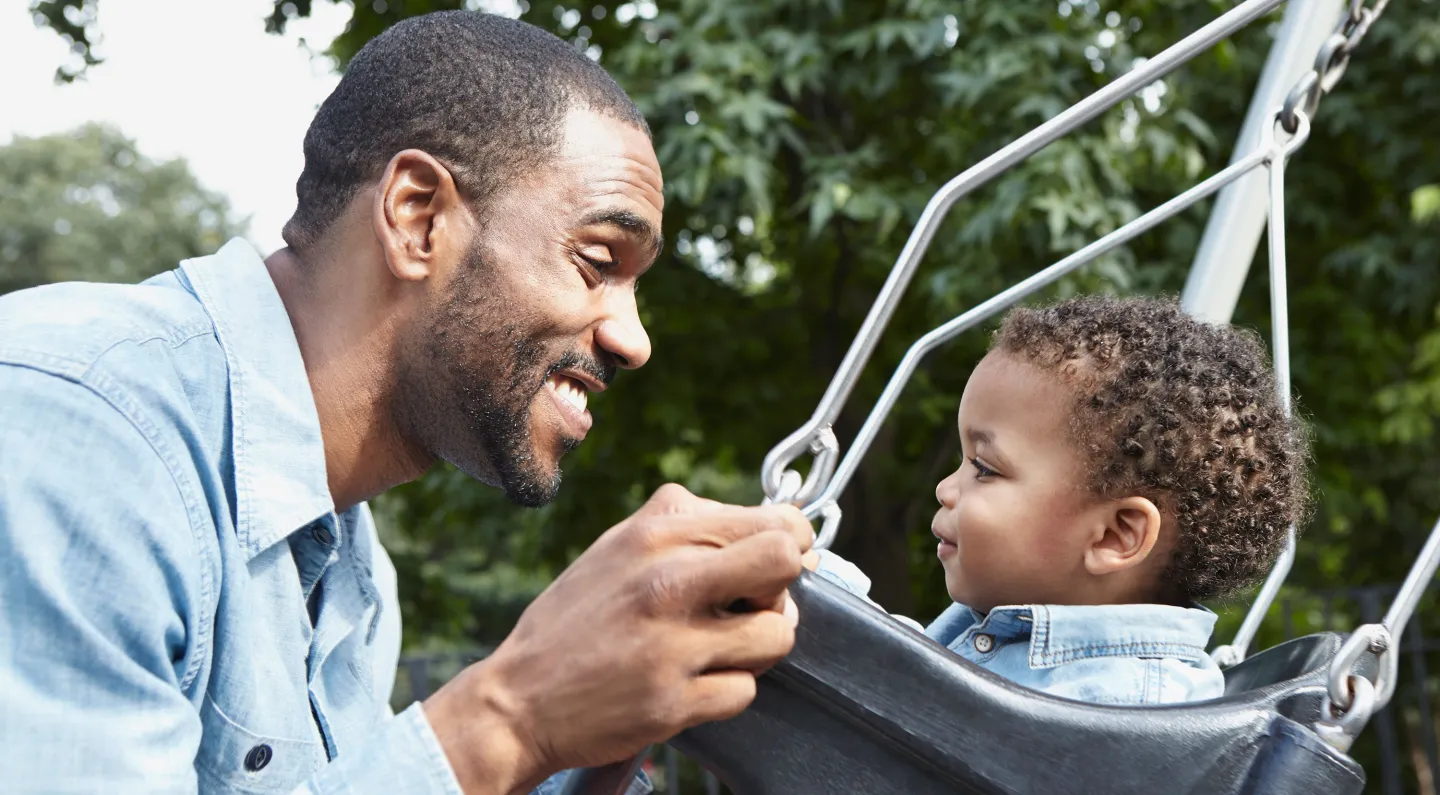 Father smiles and interacts with his young son sitting in a swing at a park.
