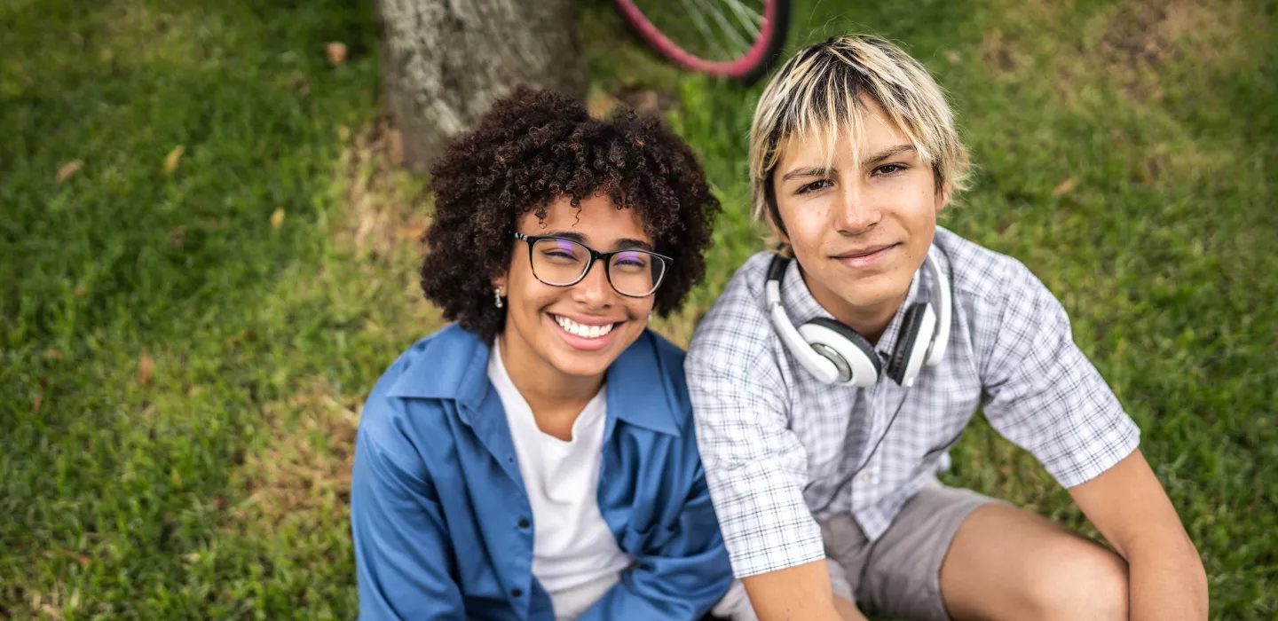 Two teens sitting on grass smiling, with a bicycle wheel visible in the background
