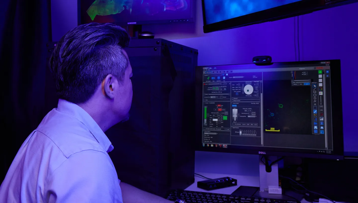 Man in lab coat examines computer screens with neural imaging data under blue lighting.