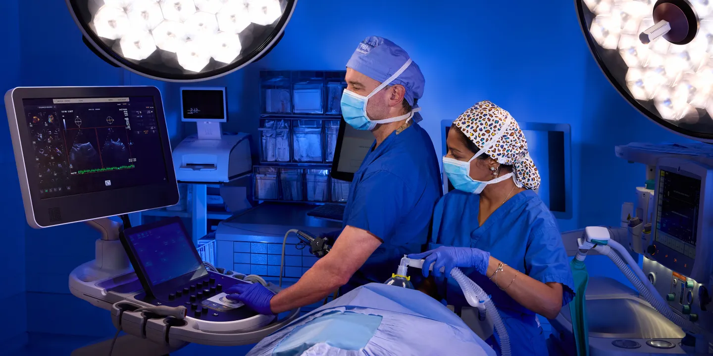 Two masked clinicians in blue scrubs operate ultrasound and anesthesia equipment in a blue-lit operating room.