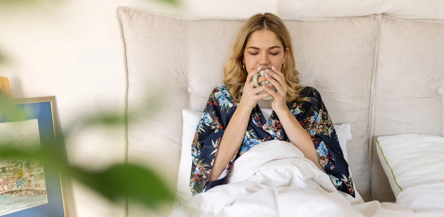 Woman relaxing in bed with eyes closed, holding a warm drink in both hands.