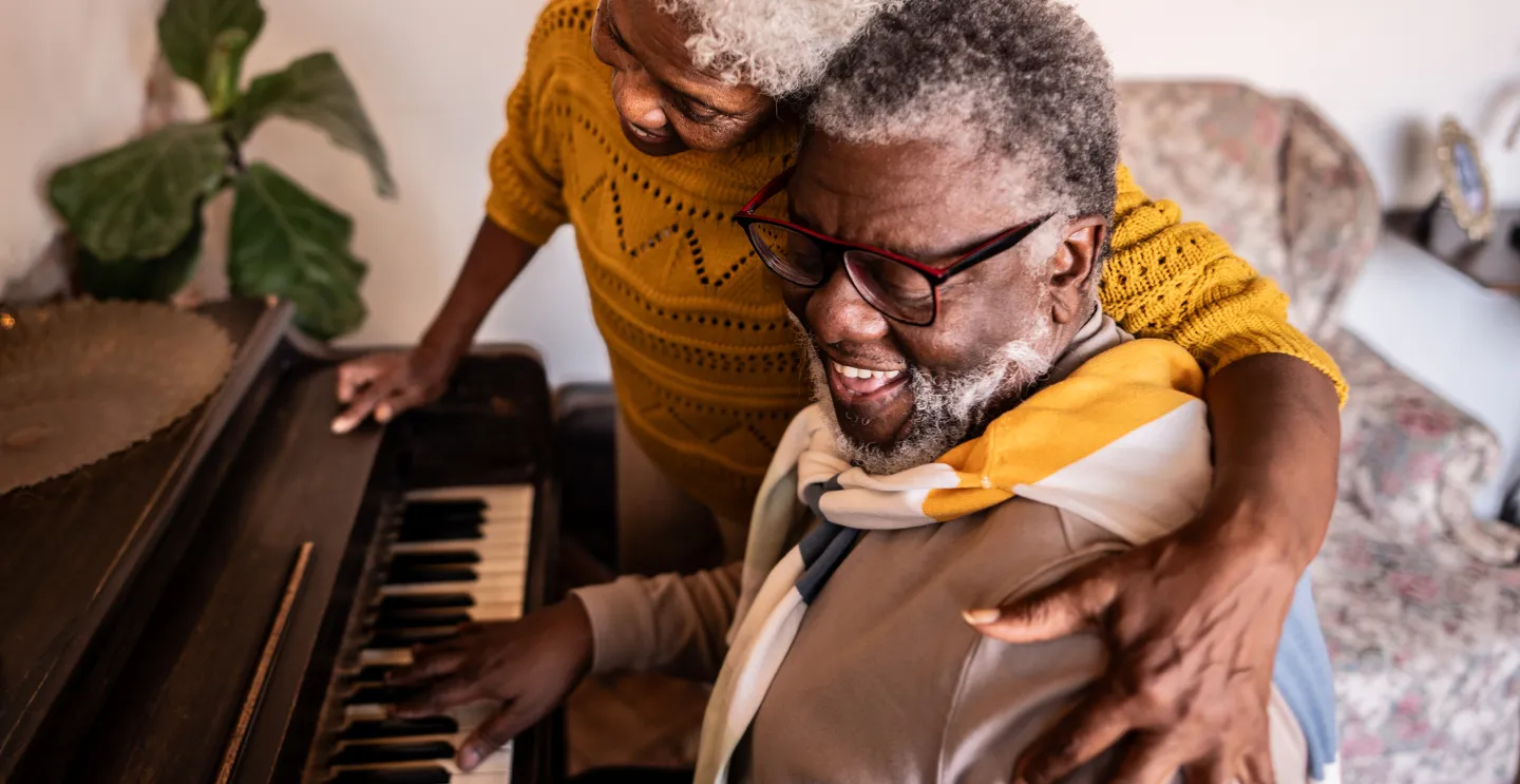 Elderly couple smiling and embracing while the man plays piano at home.