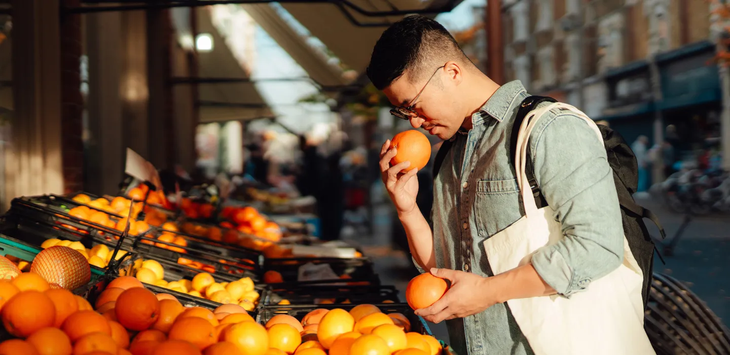 Man smelling an orange at a market stall with a tote bag over his shoulder.