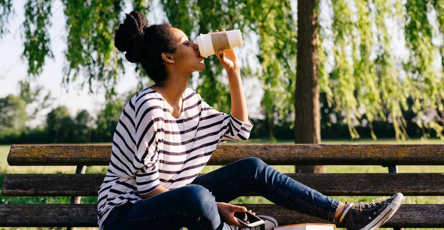 A woman in a striped shirt drinks from a disposable coffee cup while sitting on a park bench, holding a smartphone.