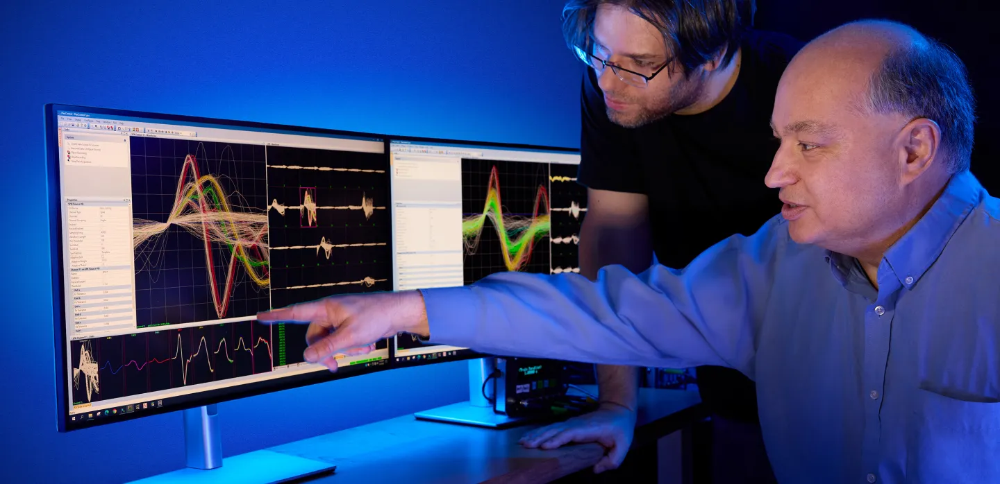 Two researchers analyzing colorful waveforms on dual monitors in a dimly lit lab with a blue light glow.