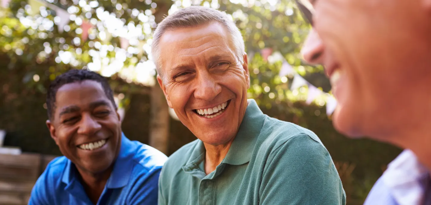 Three smiling middle-aged men sitting outdoors, laughing together in a sunlit garden setting