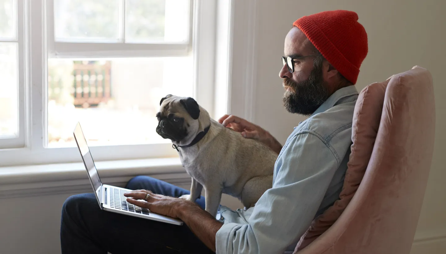Man wearing glasses and a red beanie working on a laptop with a pug sitting on his lap in a sunlit room.