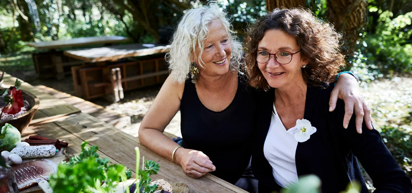 Two women sitting at an outdoor wooden table smiling and enjoying each other’s company in a lush garden setting.