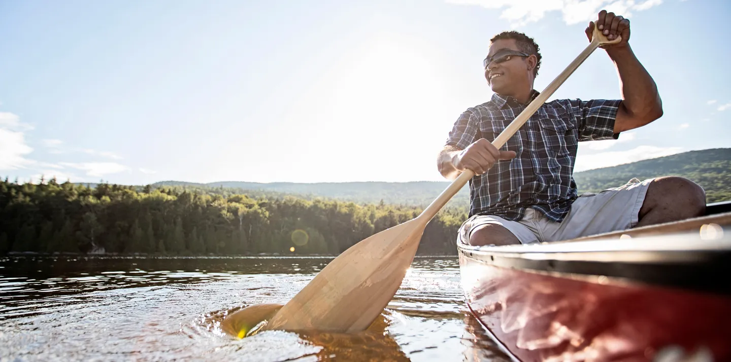 Man in a canoe paddling on a lake surrounded by forest and hills under a bright sky.