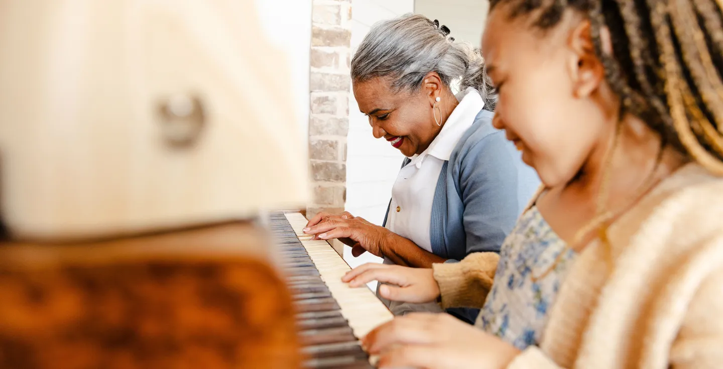 Elderly woman and young girl smiling as they play the piano together in a warmly lit room.