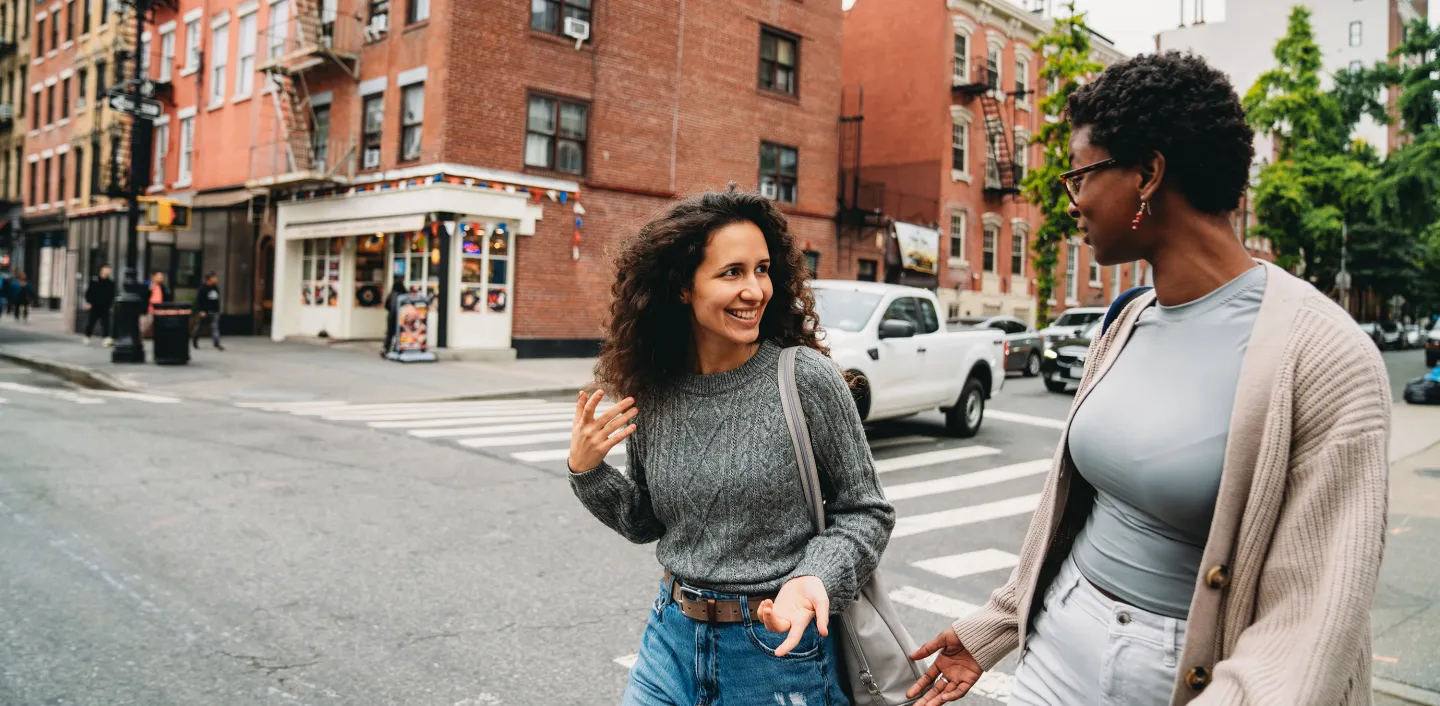 Two women walk and chat cheerfully while crossing a city street with brick buildings in the background.