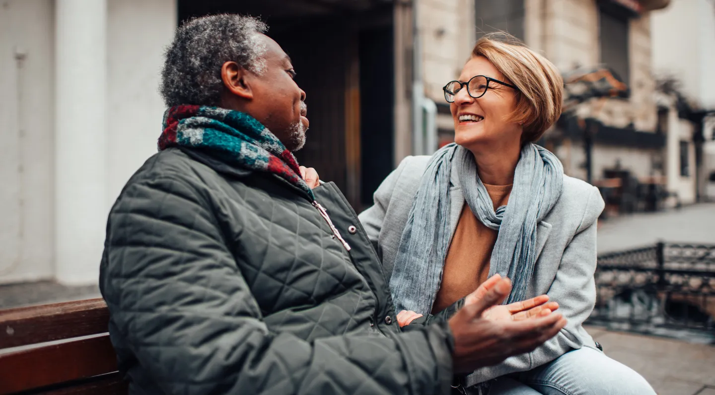 Smiling man and woman sitting on a bench outdoors, warmly dressed and engaged in a lively conversation.
