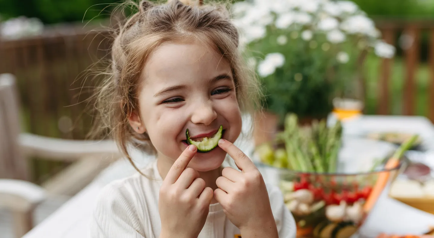 Smiling young girl holding a slice of cucumber in her mouth like a smile while sitting at an outdoor table with vegetables.