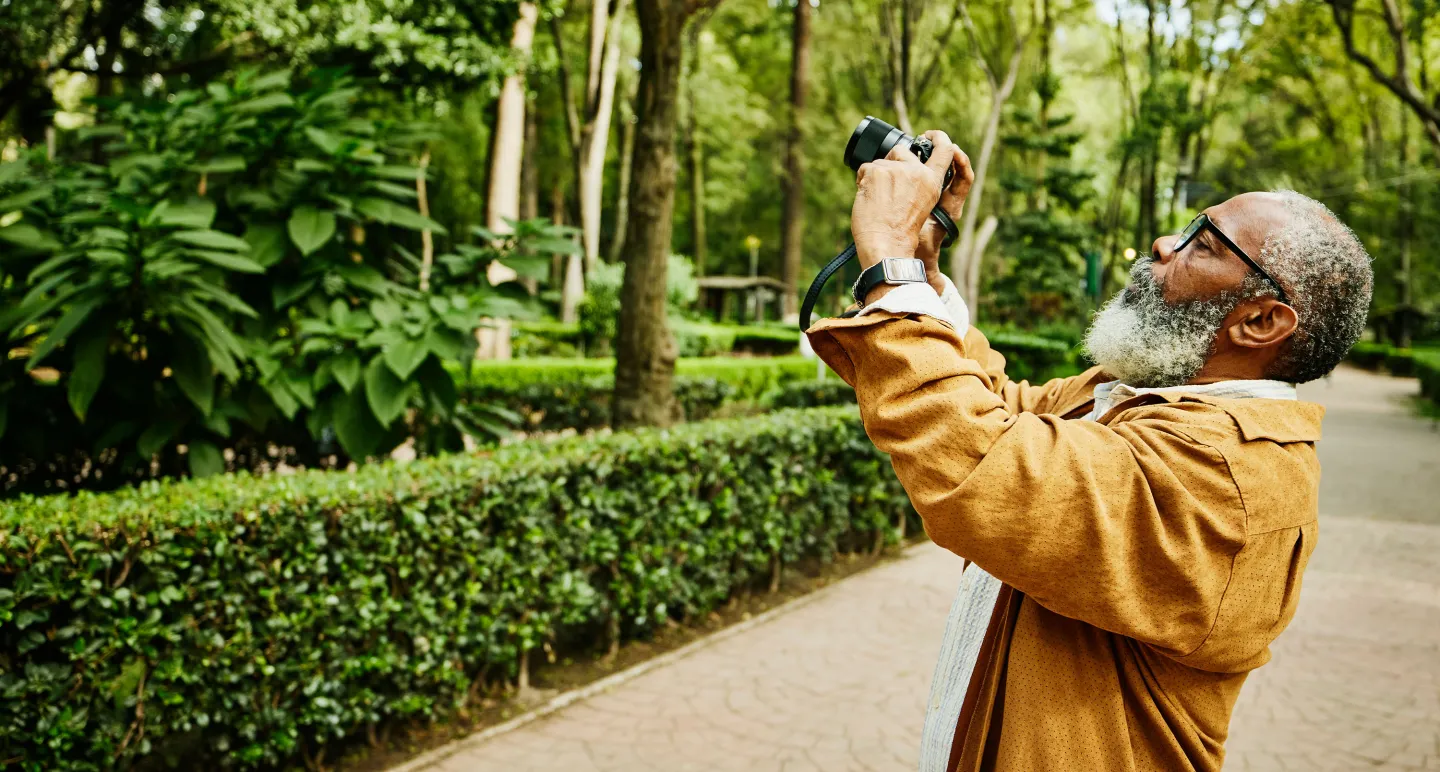 Older man with a white beard and glasses taking a photo with a camera in a lush, green park filled with trees and hedges.