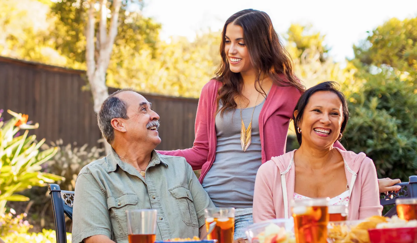 Smiling young woman standing behind two older adults seated at an outdoor table, enjoying a sunny day.
