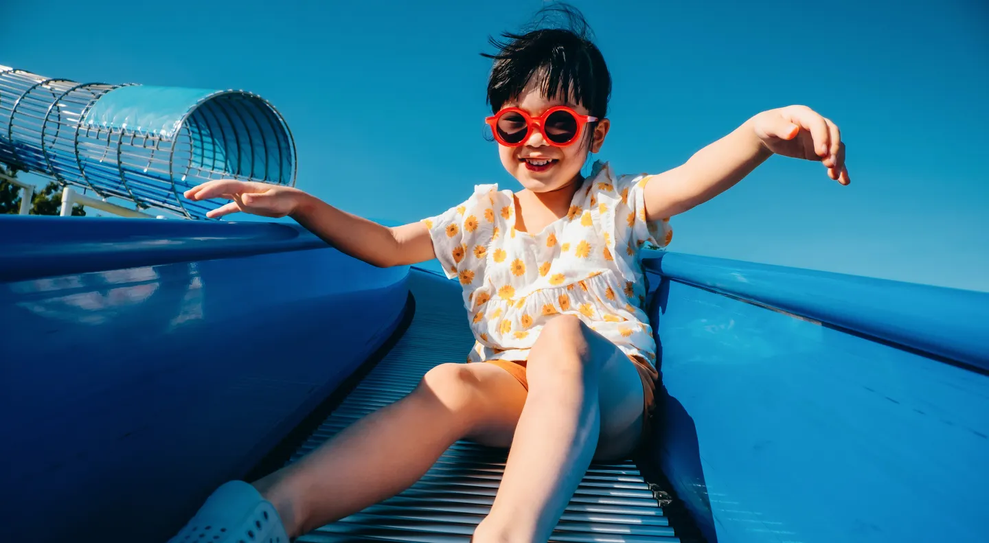 Smiling child wearing red sunglasses slides down a bright blue slide on a sunny day.