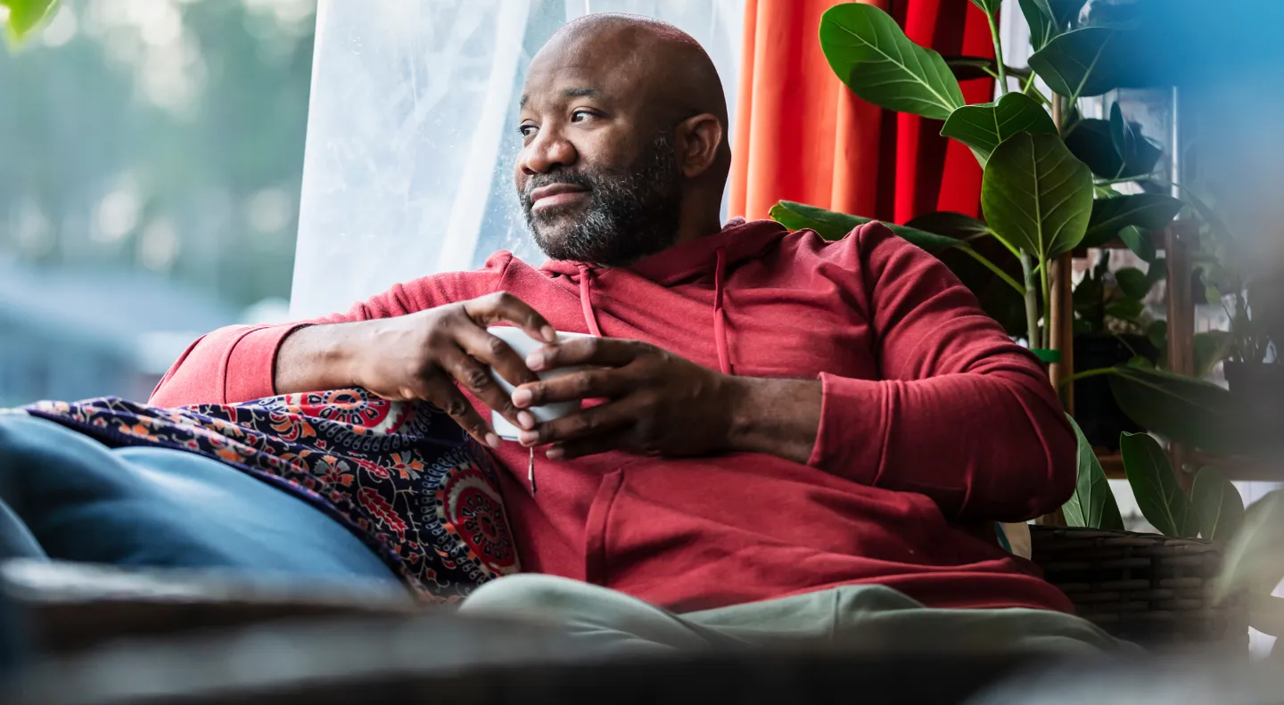 Man in a red hoodie sitting by a window, holding a mug and relaxing