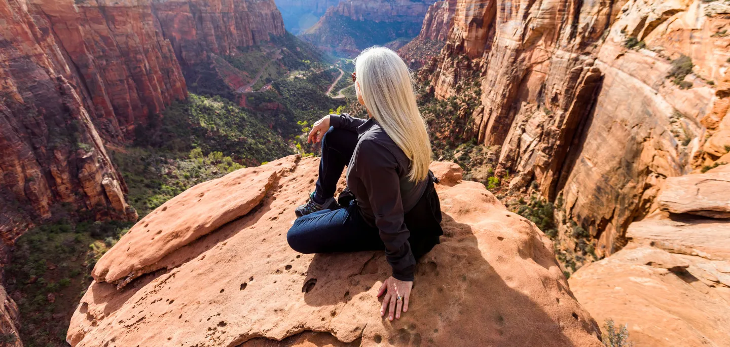 Woman with long white hair sitting on a red rock cliff, overlooking a canyon valley with winding roads and greenery.