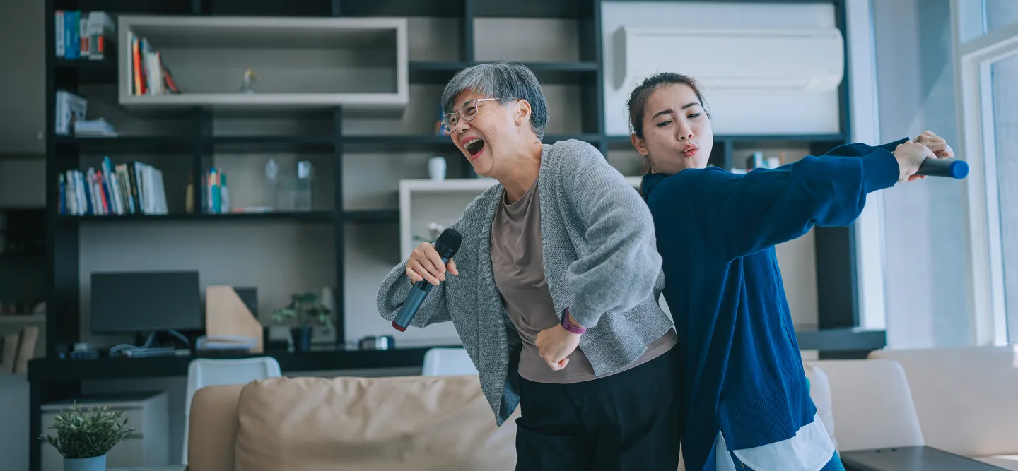 Two women laughing and singing into microphones while dancing in a cozy living room.