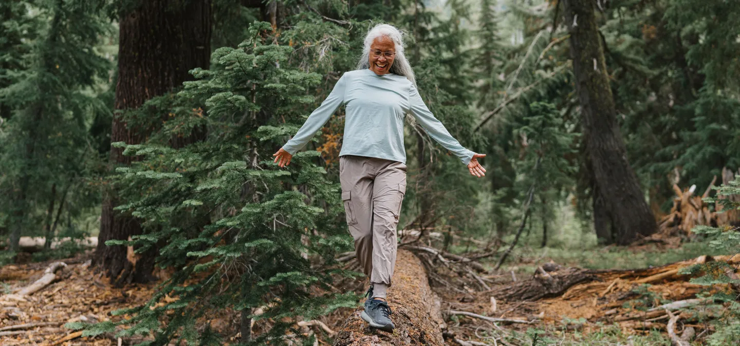 An older woman with gray hair smiles joyfully as she balances on a fallen tree trunk while hiking in a forest.