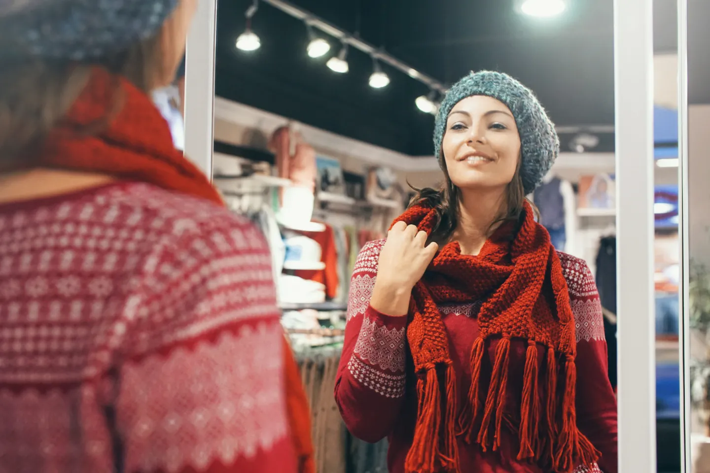 Woman smiling at herself in a mirror while wearing a knit scarf and winter hat in a clothing store.