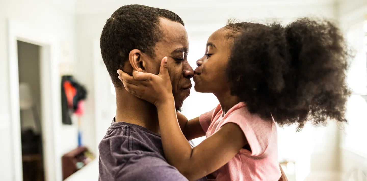 Young girl kissing her father on the nose while holding his face in a warm, sunlit room.