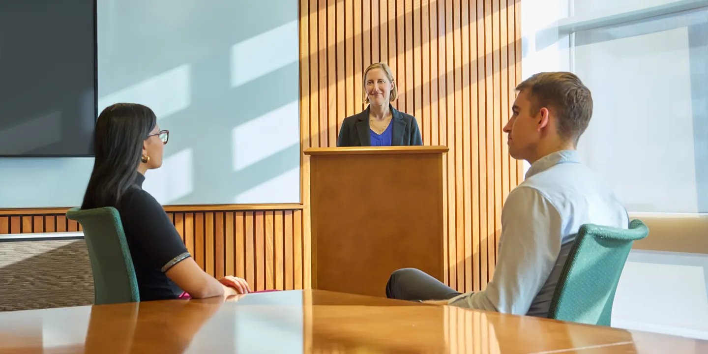 Woman speaking at a podium while two people listen attentively in a modern conference room.
